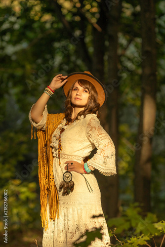A young woman strolls calmly and casually through a field; she is wearing an open yellow shawl and a brown hat, with her arms spread wide. Boho style.