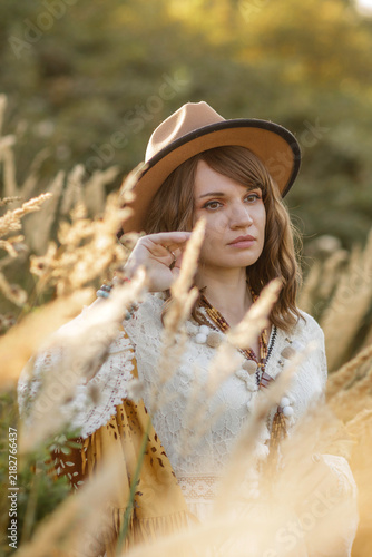 A young woman strolls calmly and casually through a field; she is wearing an open yellow shawl and a brown hat, with her arms spread wide. Boho style.