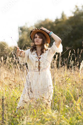A young, fair-skinned woman strolls calmly and casually through a field; she is wearing an open yellow shawl and a brown hat, with her arms spread wide. Boho style.