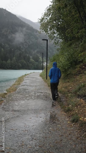 Tourist walks along a lakeside trail in the rain, enjoying the peaceful mountain scenery.