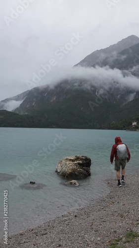Tourist climbs a large rock beside a turquoise alpine lake surrounded by rain and low clouds in a dramatic mountain landscape.