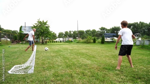 Sunny day. Two people play soccer barefoot on grass. White goal stands ready. One runs to kick ball. Other watches from distance. Casual, joyful movement