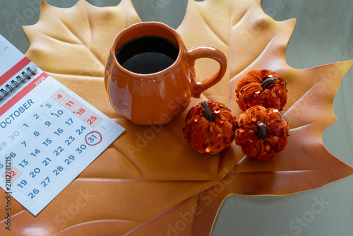 Pumpkin shaped coffee mug sitting on a decorative maple leaf with a calendar showing october 31st circled, surrounded by small glitter pumpkins, creating a cozy autumn scene