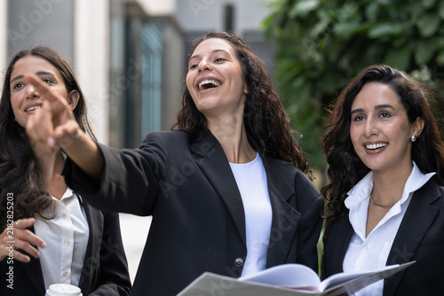 Фотография A diverse team of three Hispanic businesswomen in dark suits, wide-eyed at what she sees, look up from a report towards the skyline with a large open report held between them