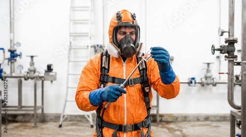 Industrial worker in full protective suit and gas mask inspecting sample in laboratory setting with pipes and valves