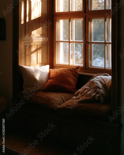 Sunlight shines through the window onto a cozy seat filled with pillows. The room has a warm, nostalgic feel with faded tones.