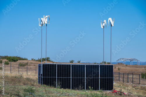 Fotografie a hybrid off-grid renewable energy system combining a dark solar panel array with four white vertical axis wind turbines in a dry rural field under a clear blue sky