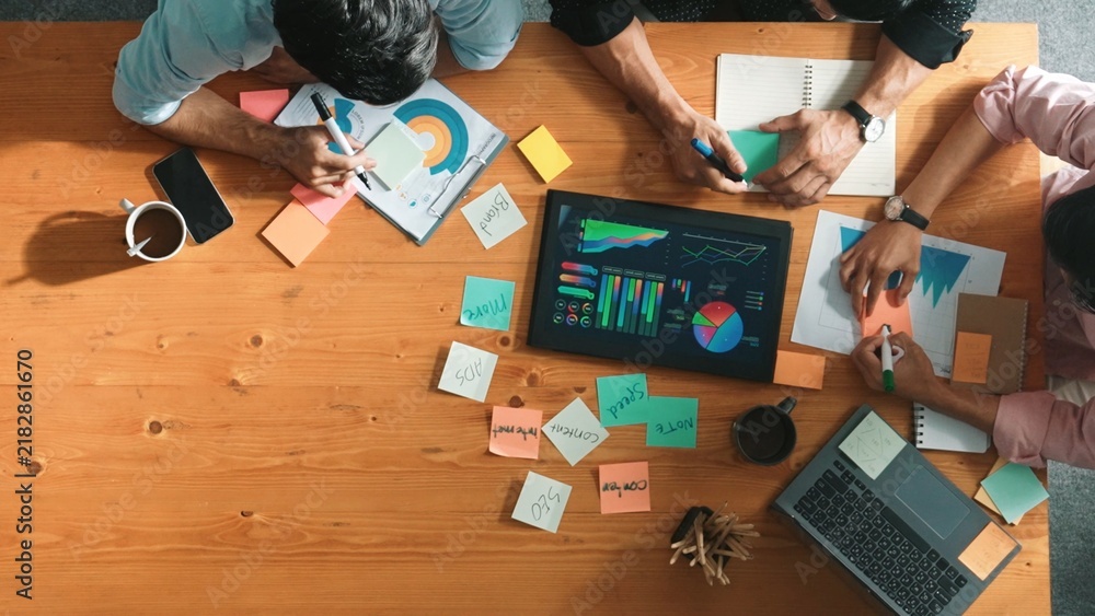 Obraz premium Top aerial view of diverse team writing colorful sticky notes to brainstorm marketing idea while looking at financial chart. Multicultural smart business people taking a note about plan. Convocation.