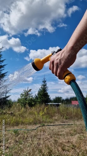 A man's hand holds a yellow watering can and pours water on a green garden against a blue sky with white clouds. A man waters plants in the yard using a garden hose with a sprayer