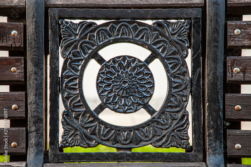Close-up view of an ornate dark wrought iron architectural detail featuring floral scrollwork, geometric patterns, vertical bars, metal rivets, and weathered textures. 