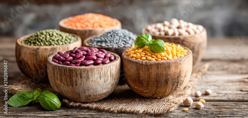 Assorted dried legumes and grains sit in rustic wooden bowls, creating warm still life with red beans, green lentils, and yellow lentils. Basil leaves add fresh color among varied ingredients.