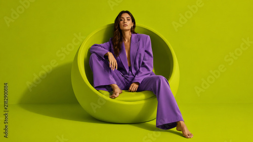 Stylish woman in a purple suit sitting in a sculptural green chair against a vibrant studio background, ideal for fashion, furniture, lifestyle and advertising campaigns.