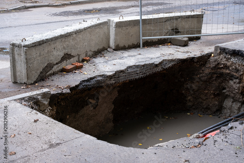 Sinkhole crater beneath sidewalk with cables and murky water, road damage close up