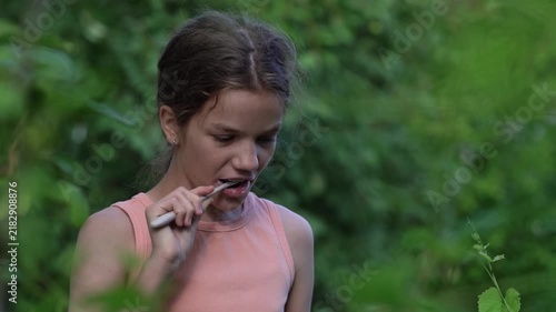 A teenage girl is brushing her teeth while outdoors, with the sky in the background, during a hiking trip.
