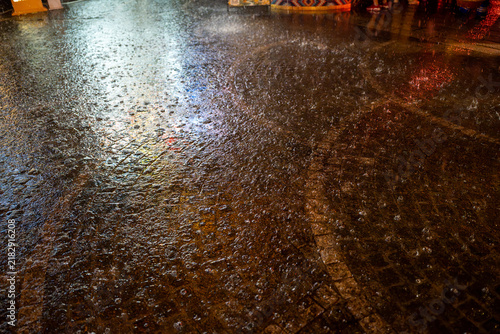 Obraz na plátně Torrential rain hitting a textured stone street at night, creating atmospheric splashes and reflecting colorful ambient city lights in the puddles