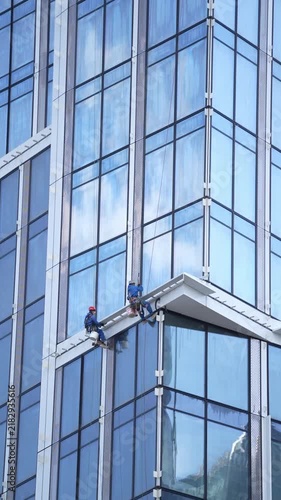 Industrial climbers suspended on ropes perform facade installation and finishing work on a modern glass high rise building, showing professional construction services and urban infrastructure