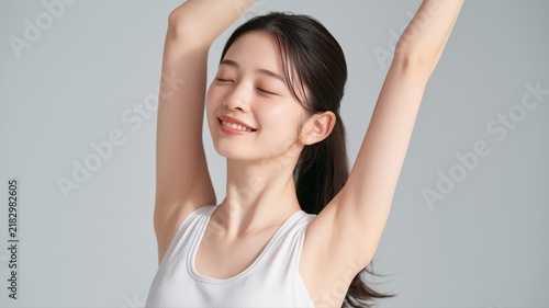 白いタンクトップを着て両腕を上げて目を閉じて微笑む若い女性。リラックスや健康的なライフスタイルのイメージ素材