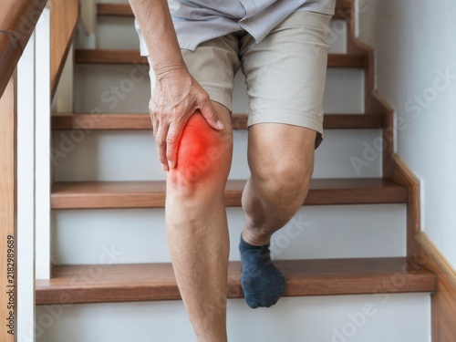 An elderly person holding their painful knee while carefully walking down a set of wooden stairs.