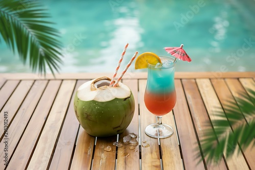 Fresh green coconut and layered tropical cocktail on a wooden table by a swimming pool side