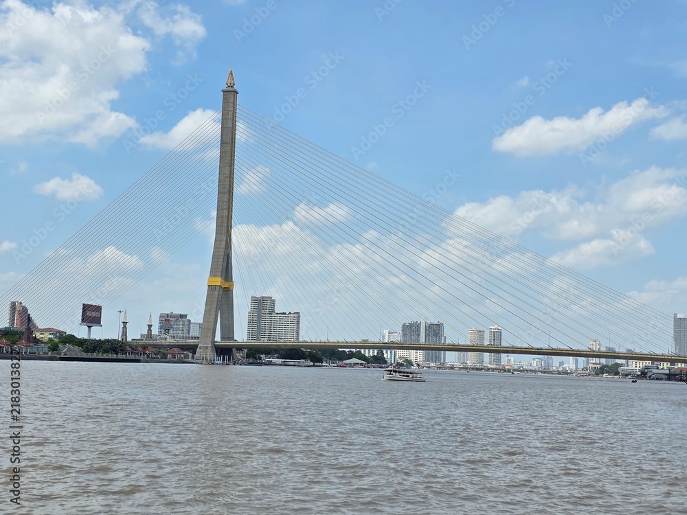 Fototapeta premium Rama VIII Bridge over Chao Phraya River: A grand cable-stayed bridge spans the wide Chao Phraya River, with modern skyscrapers forming the city skyline in the background under a partly cloudy sky.