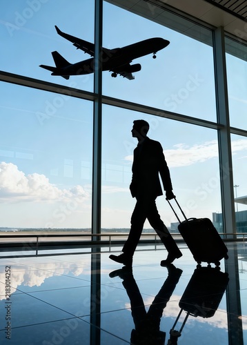 Businessman silhouette with rolling luggage walking through airport terminal, large glass windows, airplane flying outside, bright blue sky, highly reflective floor — corporate travel concept.