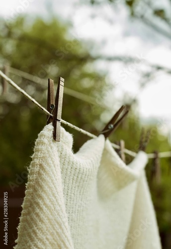Close up side view of a white knitted sweater hanging on an outdoor clothesline held by rustic wooden clothespins.