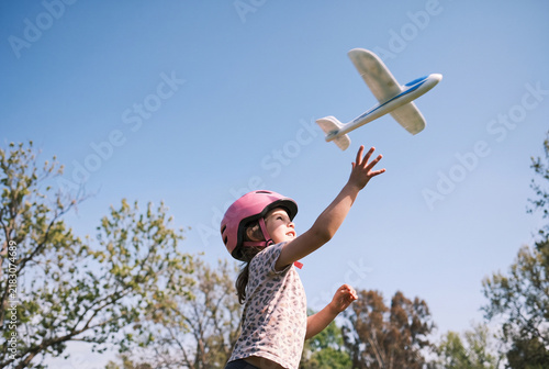 Cheerful Little Girl in Pink Helmet Throws Model Airplane into the Sky in a Sunny Park, Outdoor Play and Active Childhood