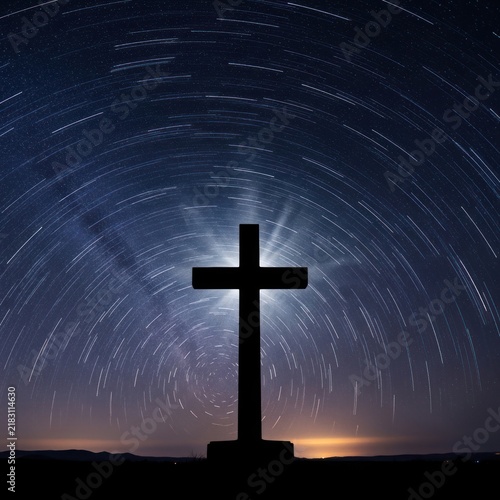 A dark silhouette of a large wooden cross stands prominently against a breathtaking night sky filled with swirling star trails.
