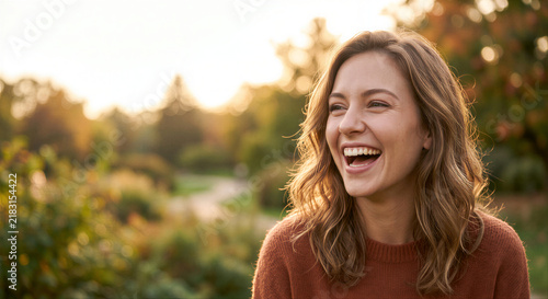 Cheerful young woman laughing while standing outdoors in a sunny autumn park during golden hour