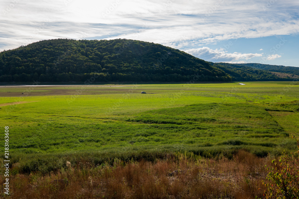Obraz premium Car driving across the exposed grassy bed of Domaša reservoir in Slovakia, with the Ondava River flowing through the drained landscape during extremely low water levels.