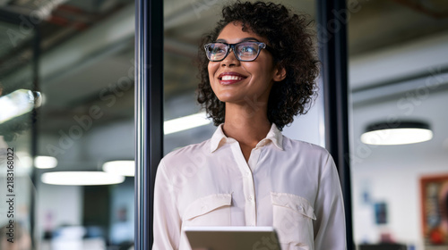 Happy confident young African American professional woman, female entrepreneur, businesswoman looking away holding tab standing in office at work dreaming of future success