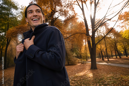 Smiling young man wearing a navy coat in an autumn park. Vertical portrait of a happy male enjoying a sunny fall day outdoors. Seasonal fashion and lifestyle concept