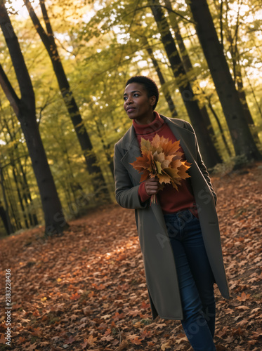 Black woman holding a bouquet of autumn leaves in a forest. Vertical portrait of a female walking in the woods during fall. Seasonal outdoor lifestyle and nature concept