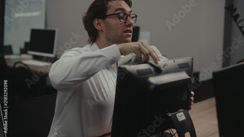 Young businessman in glasses wiping down computer monitor with cloth sitting at desk. Office worker male tidying up workspace for hygiene and cleanliness. Shooting in slow motion.
