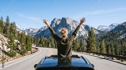 Woman standing through car sunroof with arms raised on mountain road.