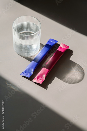 Glass of water with colorful powder drink stick sachets on table with sunlight and shadow.