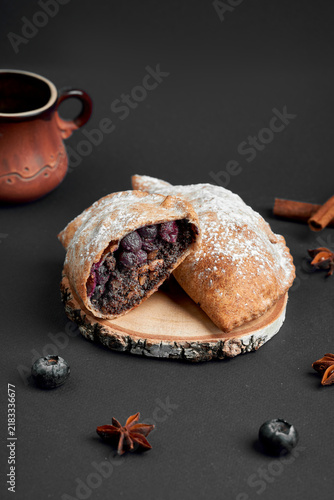Halved sweet baked pie with poppy seed and blueberry filling dusted with powdered sugar on wood slice. Dessert pastry on dark background.