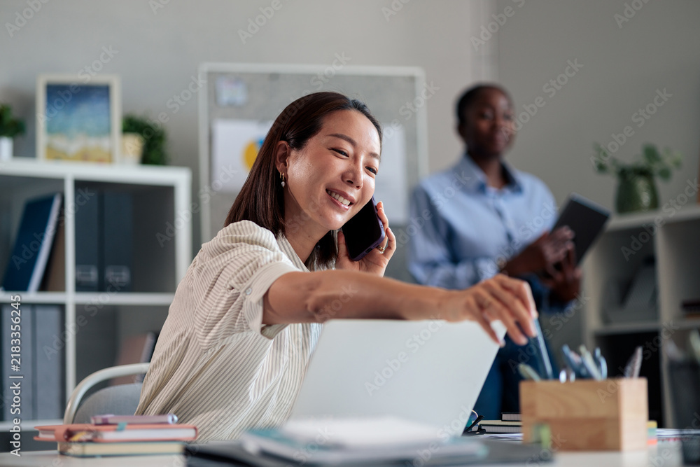 Fototapeta premium Asian businesswoman smiling and talking on smartphone in office