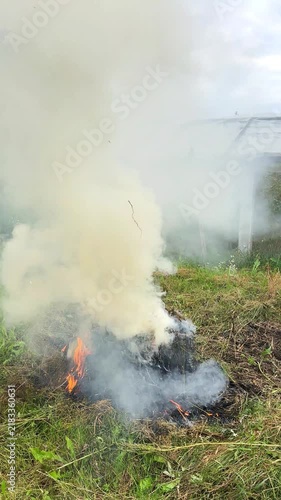 A smoldering pile of dry grass and brush burns on the ground, producing heavy white-gray smoke in a grassy area outdoors, highlighting fire danger and air pollution concerns. Vertical video background