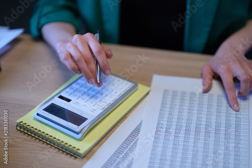 Woman accountant audits data in spreadsheet and counts on digital calculator at desk. Female inspector monitors company expenses with pen in office