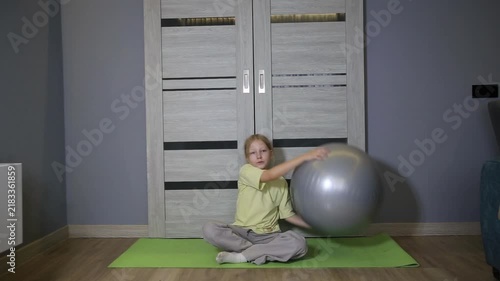 Young blonde girl sitting on yoga mat with fitness ball indoors, resting during home exercise routine, promoting healthy lifestyle for children, physical activity at residence