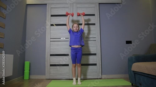 Young caucasian girl performing shoulder press exercise with red dumbbells indoors at home, standing on a yoga mat, developing strength and fitness during day.