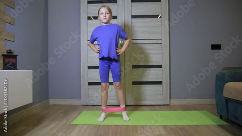 Young girl performing side steps with resistance band during home fitness workout. Child exercising indoors, focusing on lower body strength and movement, active lifestyle.