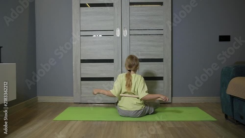Little girl stretching back with wooden stick on yoga mat indoors. Young caucasian female child engaging in physical exercise at home, practicing flexibility and fitness routine.