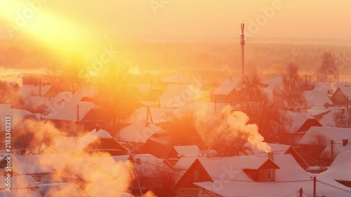 Aerial view winter village landscape frosty morning golden sunlight rising smoke chimney tops snow covered rooftops communication tower background cold weather scene peaceful rural settlement