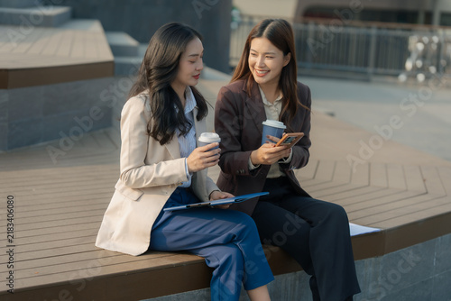 Two Asian businesswomen discussing work ideas over coffee with laptop and papers on wooden table.