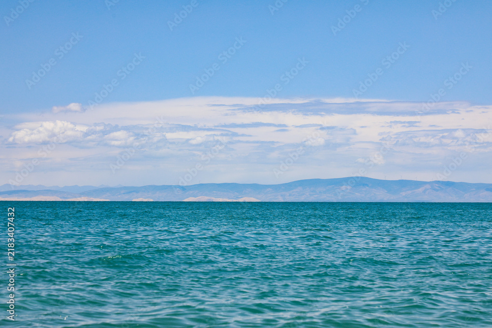 Fototapeta premium Calm blue lake with distant mountains under a bright cloudy sky
