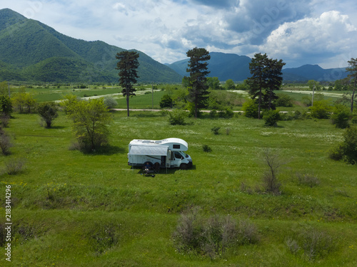 White camper van enjoying an outdoor camping trip, parked in a lush green field under a clear sky, surrounded by rolling mountains and trees, embodying freedom and travel