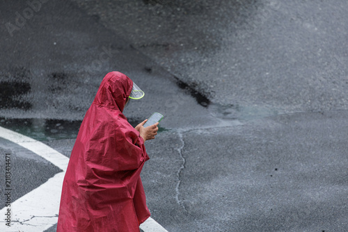 Pedestrian standing alone on a wet urban street corner wearing a hooded maroon rain poncho with a clear visor and holding a smartphone with both hands.