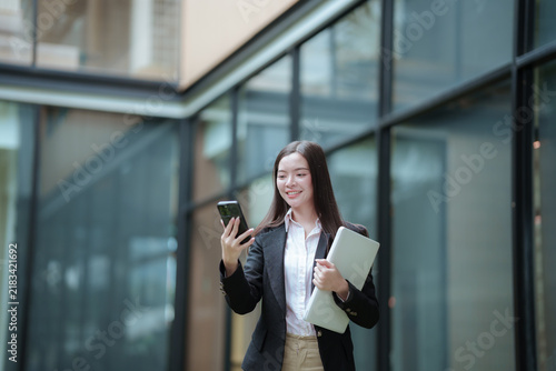 Confident Asian businesswoman using a smartphone and carrying a laptop in a modern office, representing digital communication, technology, and freelance career.